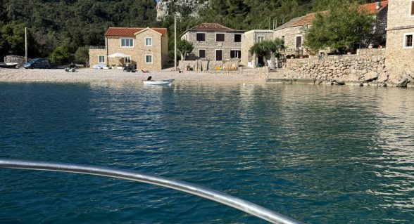 View from a boat approaching a small stone village on the Adriatic coast, with turquoise water and steep cliffs in the background