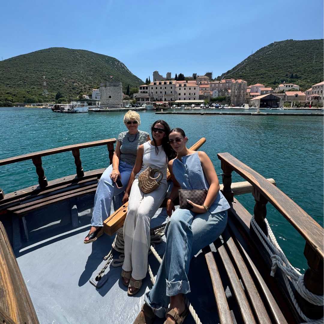 Three women sitting on a wooden boat, smiling with the historic town of Mali Ston and its medieval walls in the background on a sunny day Three women sitting on a wooden boat, smiling with the historic town of Mali Ston and its medieval walls in the background on a sunny day