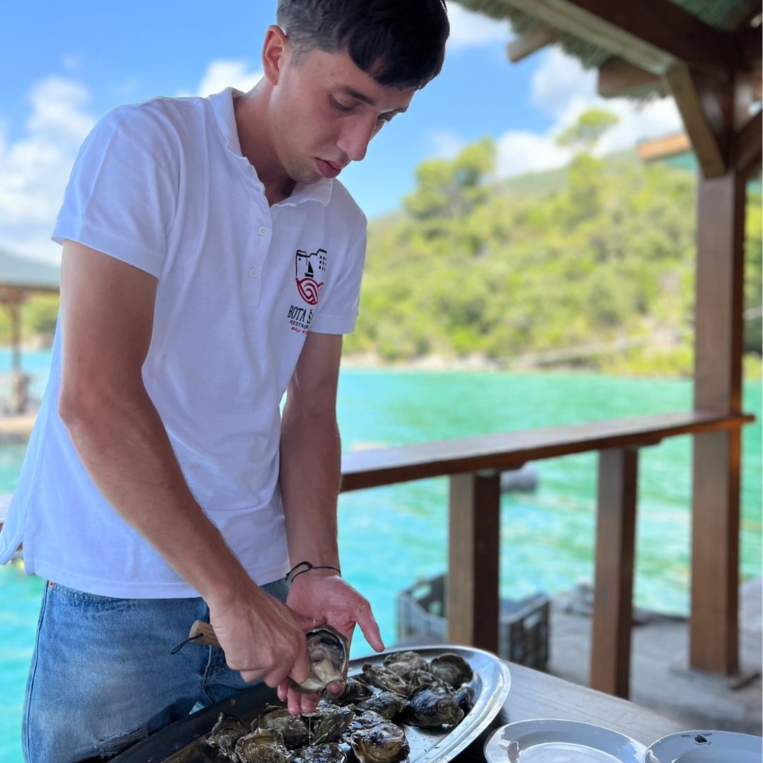 A young man shucking oysters at a wooden table overlooking turquoise water in Mali Ston A young man shucking oysters at a wooden table overlooking turquoise water in Mali Ston