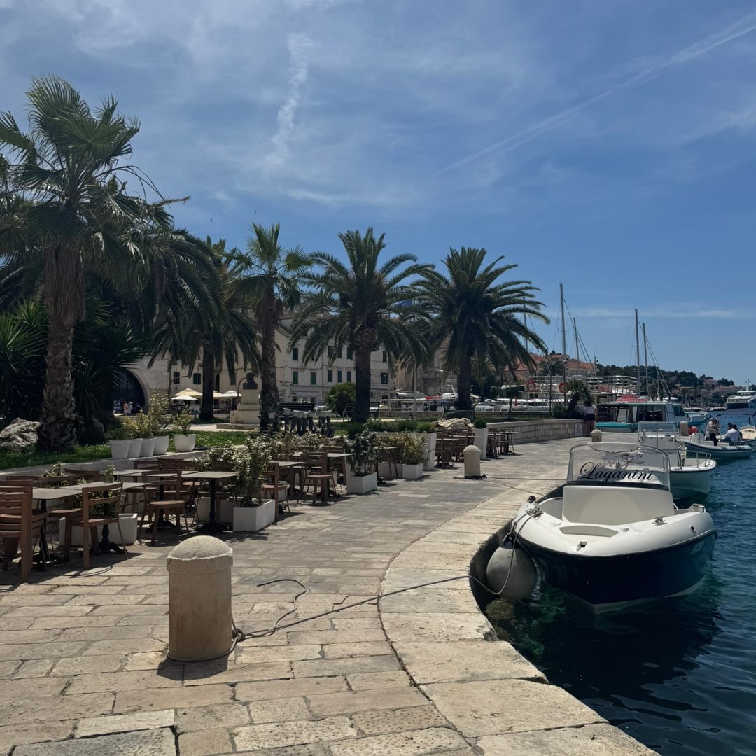 Palm-lined promenade in Hvar Town with outdoor caf&eacute;s, boats moored along the waterfront, and a bright summer sky.