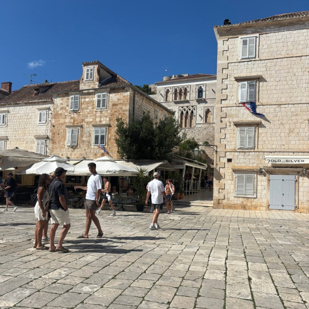 Tourists walking across the stone-paved main square in Hvar Town, surrounded by historic Venetian buildings under a clear blue sky.