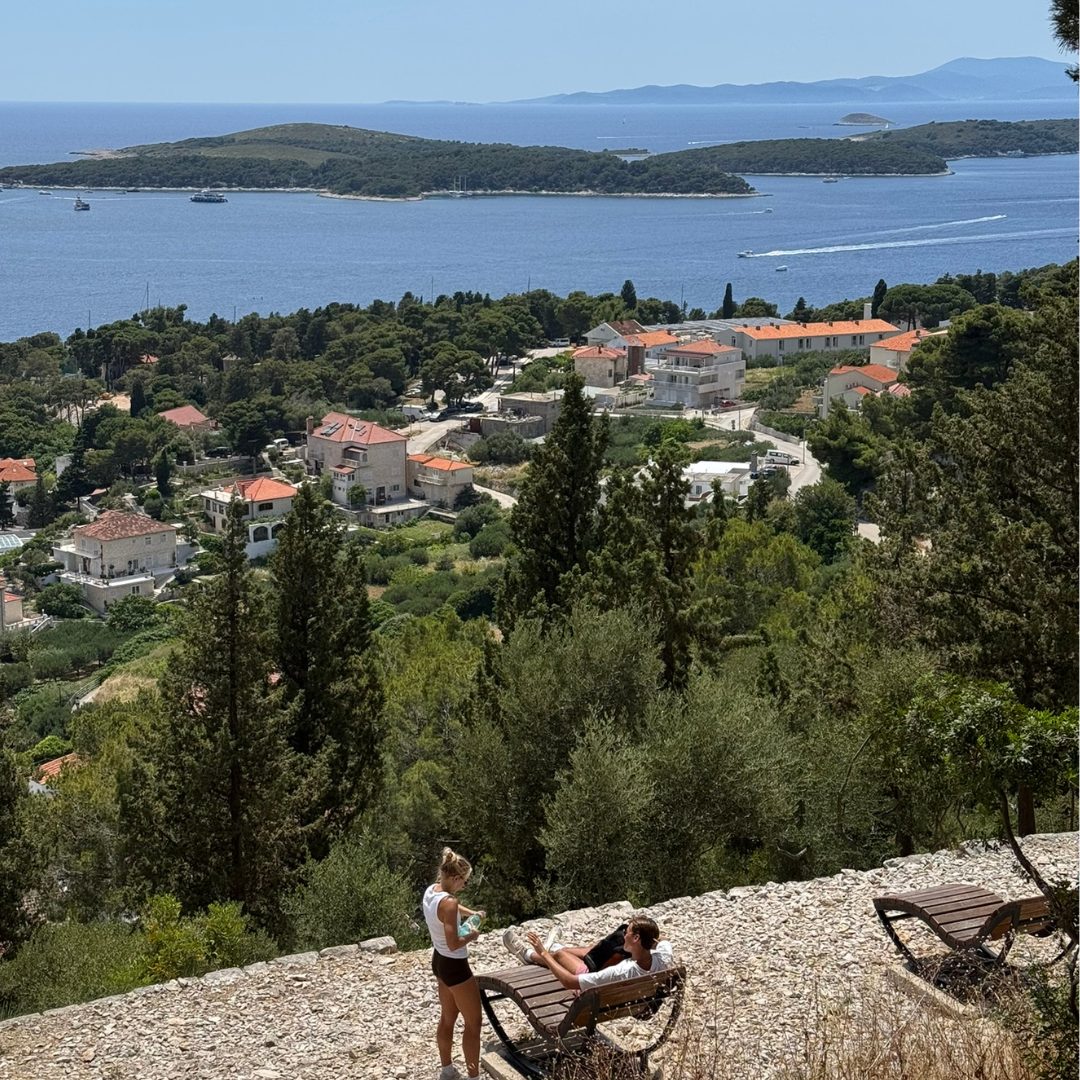 Panoramic view from above Hvar Town with people relaxing on benches, overlooking the Adriatic Sea and the Pakleni Islands.