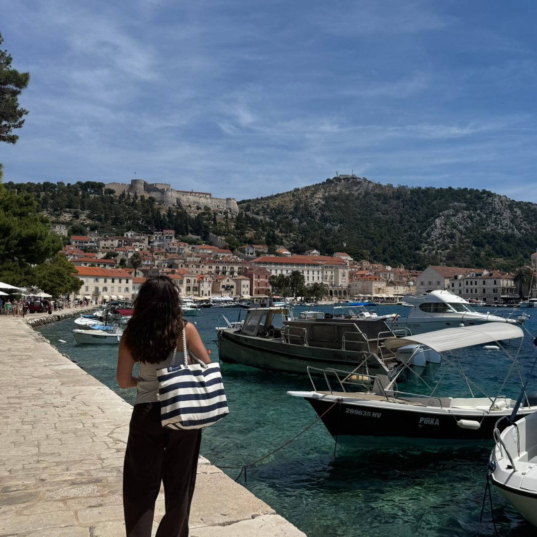 Woman walking along the Hvar Town seaside promenade with boats in the harbour and the Spanish Fortress overlooking the town from the hilltop