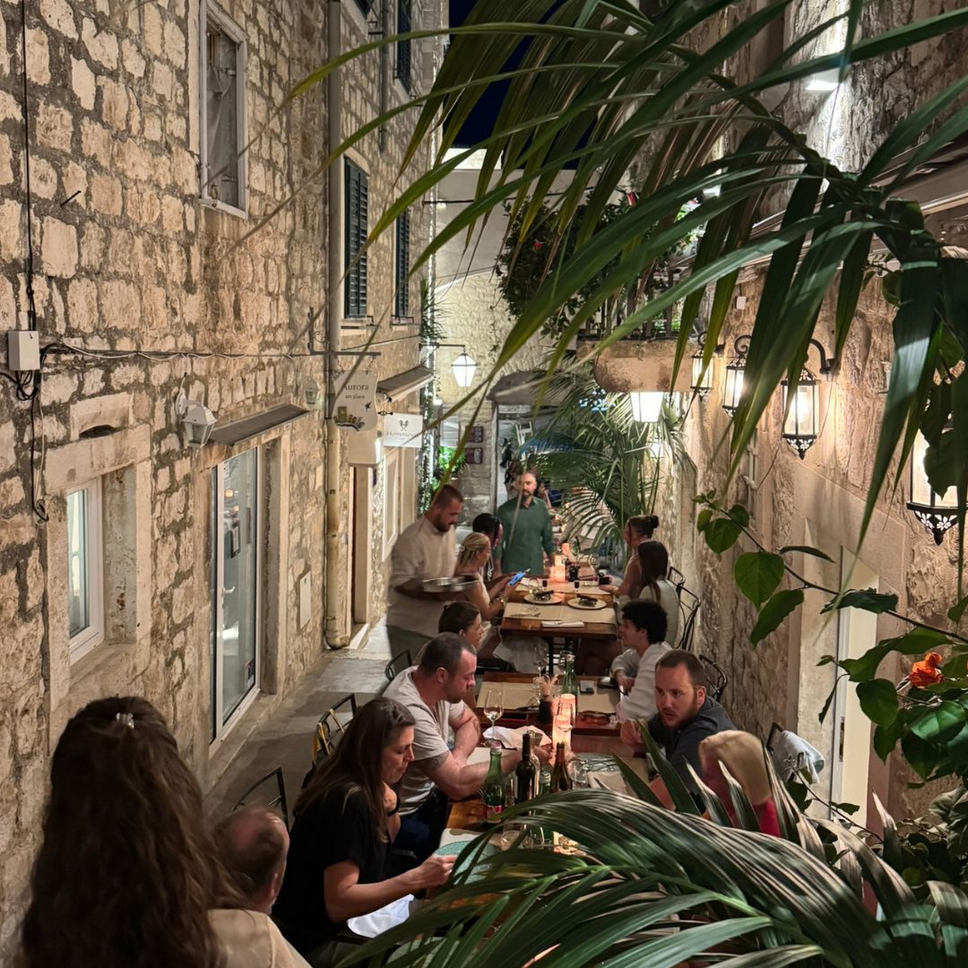 Evening dining scene in a narrow Hvar alley, with stone walls, palm leaves, and guests enjoying dinner at candlelit tables.