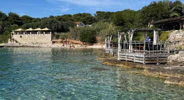 Crystal-clear turquoise waters at a beach on the Pakleni Islands near Hvar, with stone terraces and shaded lounge areas.