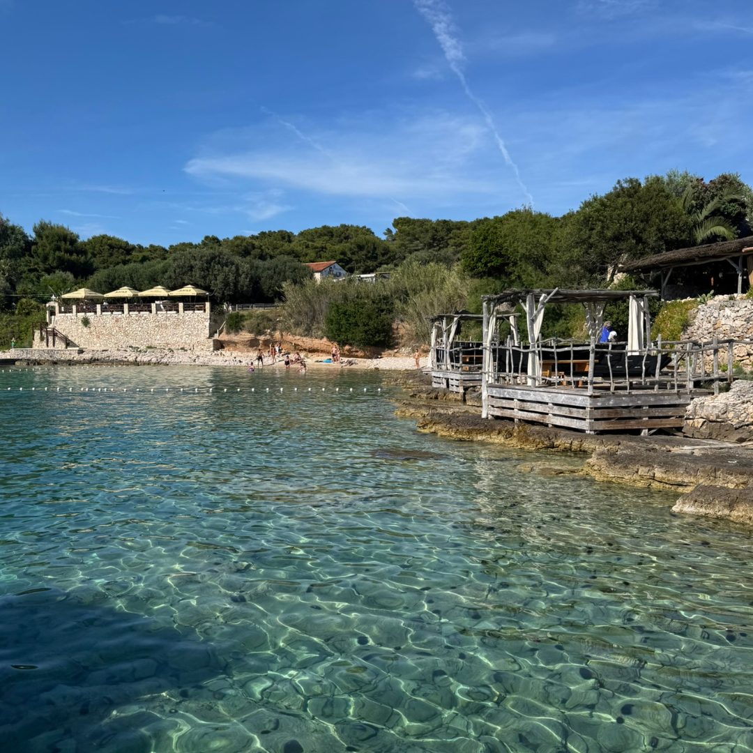 Crystal-clear turquoise waters at a beach on the Pakleni Islands near Hvar, with stone terraces and shaded lounge areas.