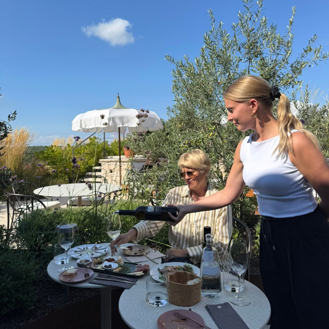 Woman pouring wine for a guest seated under a white umbrella at Radovan Wines terrace. Woman pouring wine for a guest seated under a white umbrella at Radovan Wines terrace.