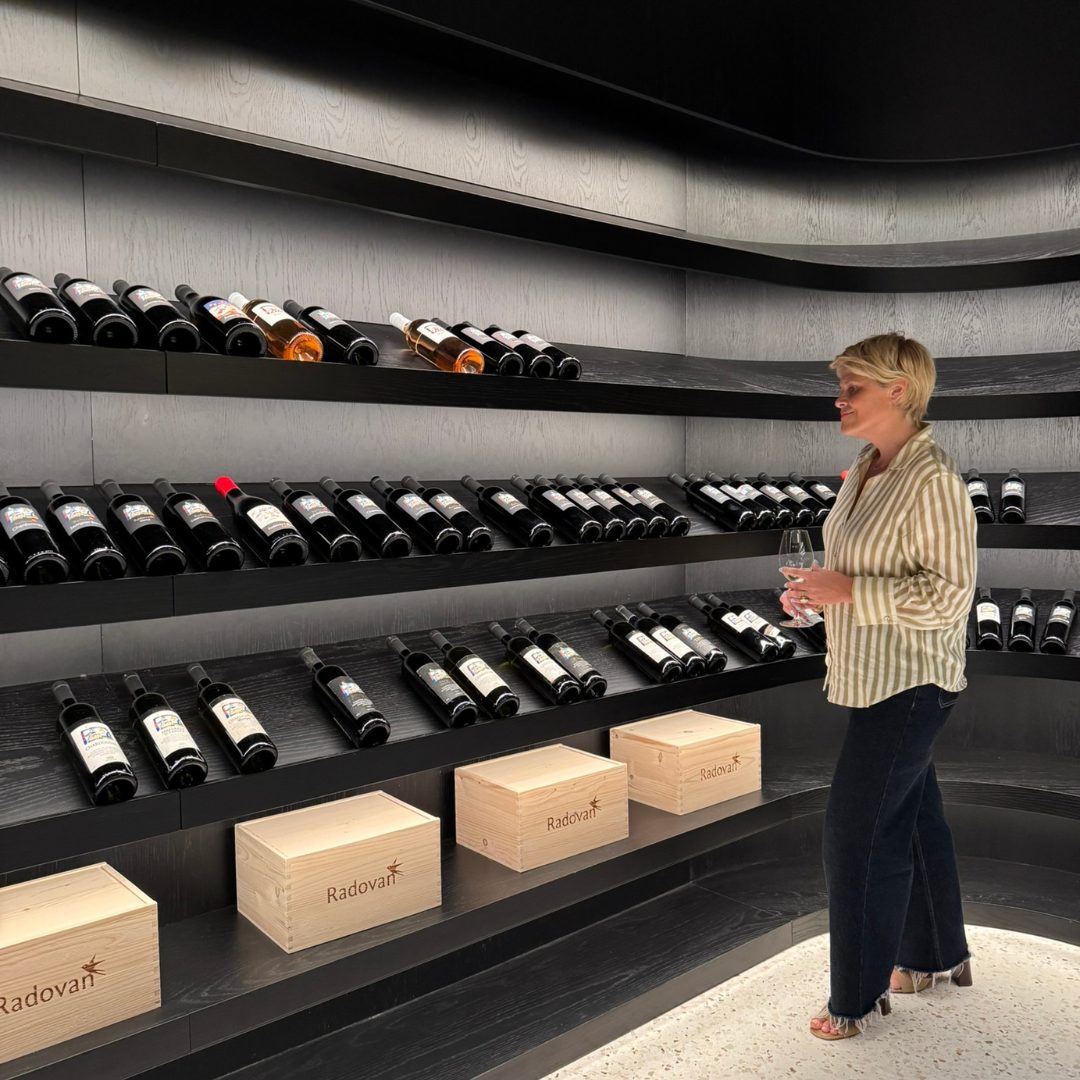 Woman holding a wine glass while browsing shelves filled with Radovan Wines bottles in the modern winery