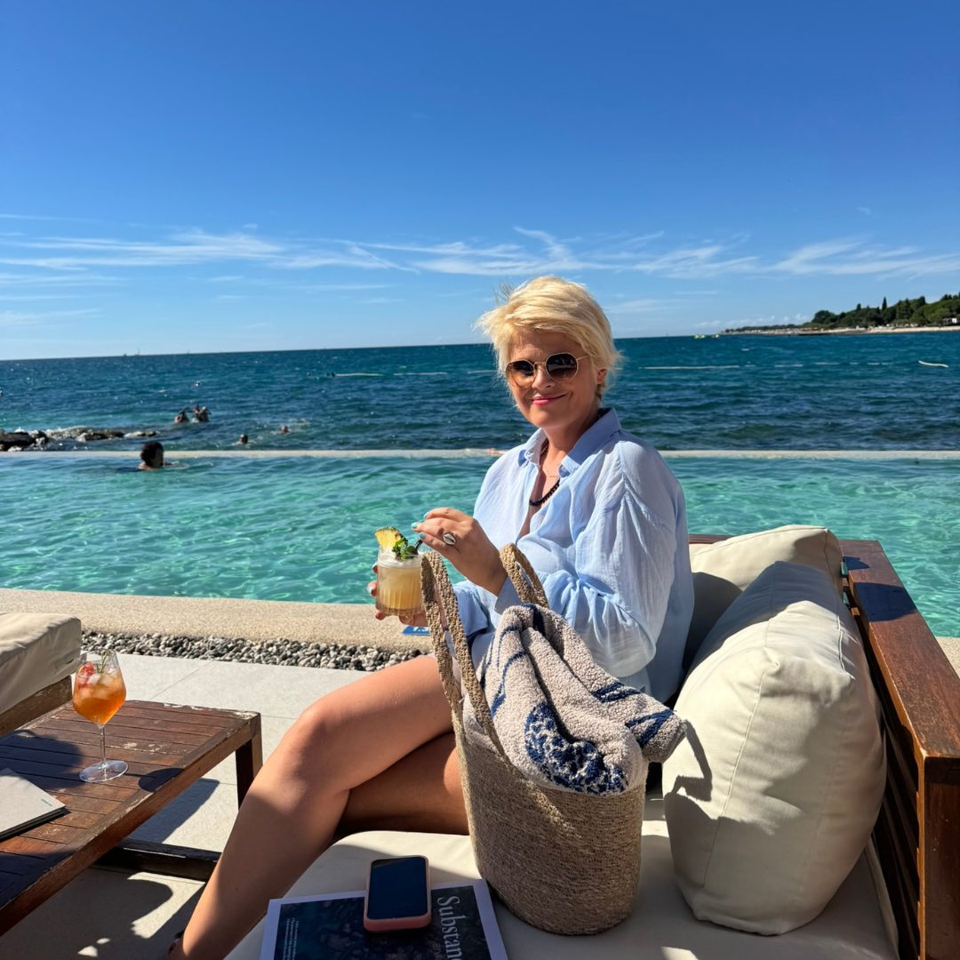 A person sitting on a lounge chair by the pool, holding a cocktail and smiling, with a beach bag, magazine, and phone on the side table. A person sitting on a lounge chair by the pool, holding a cocktail and smiling, with a beach bag, magazine, and phone on the side table.