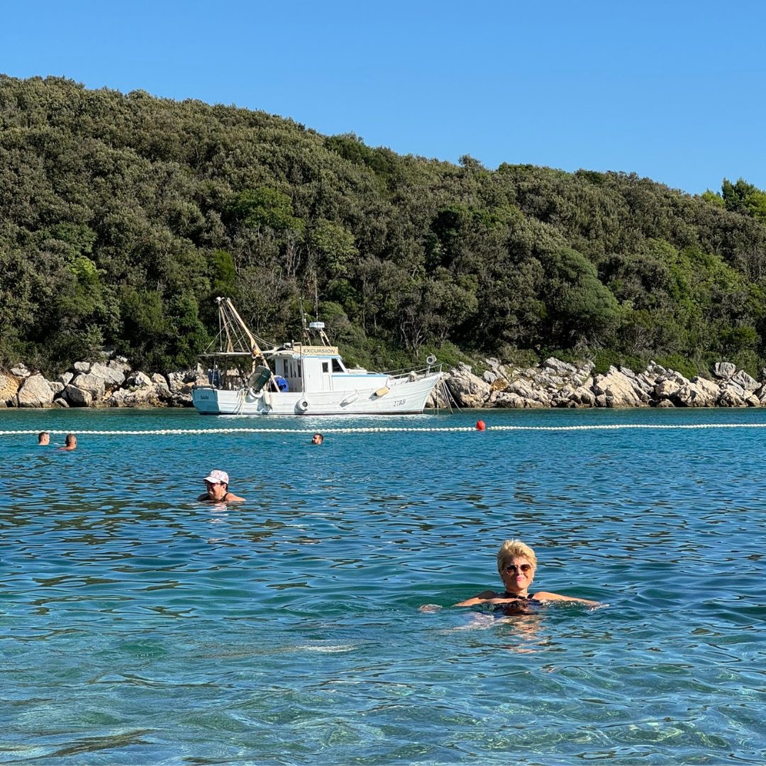 People swimming in the clear blue sea near Rab Island, with a white boat anchored by the forested coast. People swimming in the clear blue sea near Rab Island, with a white boat anchored by the forested coast.