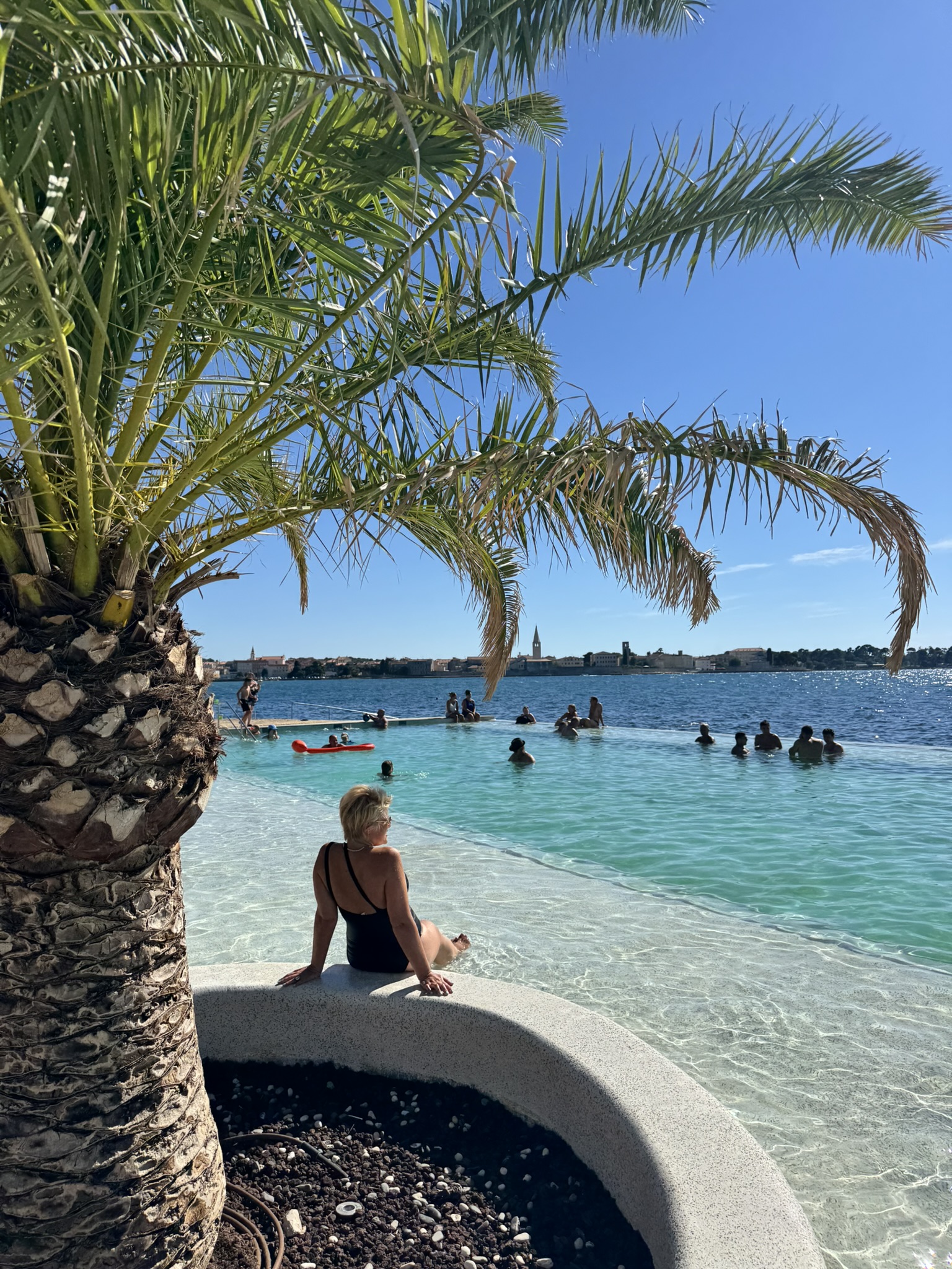 A woman enjoying the seaside infinity pool lined overlooking the calm blue Adriatic sea and Poreč old town in the distance. A woman enjoying the seaside infinity pool lined overlooking the calm blue Adriatic sea and Poreč old town in the distance.
