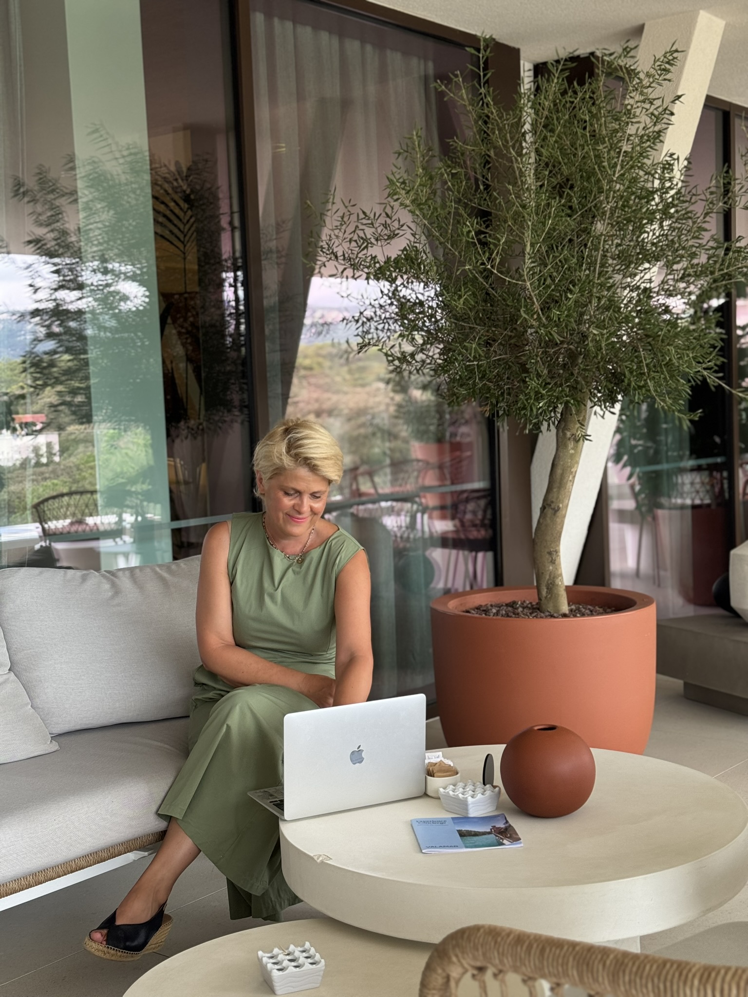 Woman working on a laptop at Arba Resort terrace, surrounded by Mediterranean-inspired decor and an olive tree in a terracotta pot. Woman working on a laptop at Arba Resort terrace, surrounded by Mediterranean-inspired decor and an olive tree in a terracotta pot.