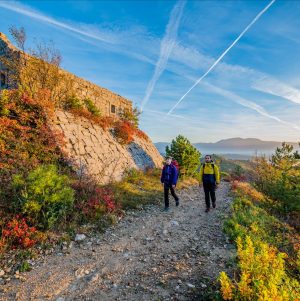 Two hikers walking along a trail beside an old stone building on a hillside, surrounded by autumn-coloured vegetation under a bright blue sky.