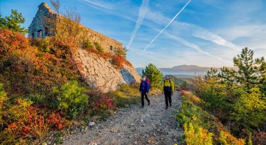 Zwei Wanderer gehen einen Pfad entlang, der an einem alten Steingebäude an einem Hang vorbeiführt, umgeben von herbstlicher Vegetation unter strahlend blauem Himmel.