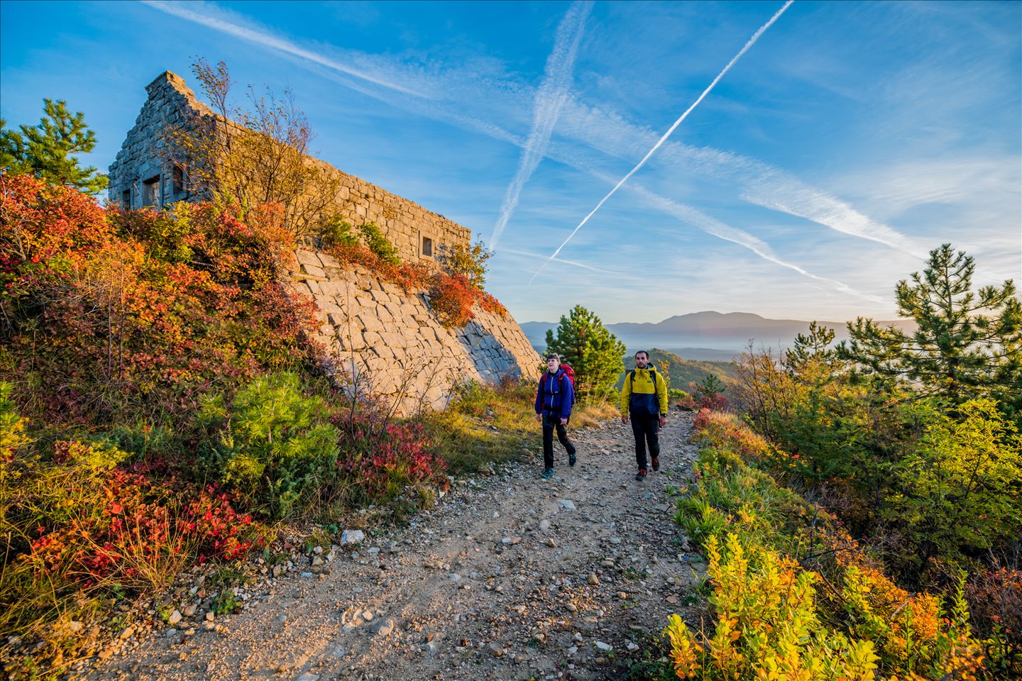 Two hikers walking along a trail beside an old stone building on a hillside, surrounded by autumn-coloured vegetation under a bright blue sky. Two hikers walking along a trail beside an old stone building on a hillside, surrounded by autumn-coloured vegetation under a bright blue sky.