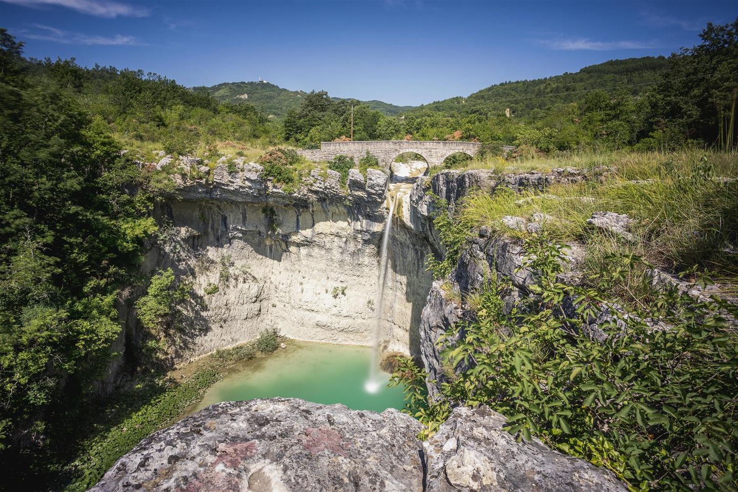 A scenic stone bridge spanning a deep rocky gorge with a narrow waterfall dropping into a turquoise pool, surrounded by lush green hills under a clear blue sky. A scenic stone bridge spanning a deep rocky gorge with a narrow waterfall dropping into a turquoise pool, surrounded by lush green hills under a clear blue sky.
