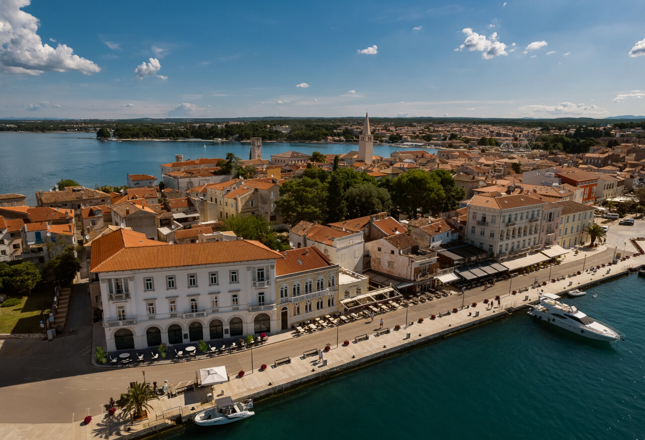 Aerial view of Poreč old town waterfront with the Jadran Heritage Hotel on the promenade, terracotta rooftops, church towers, and boats on the Adriatic.