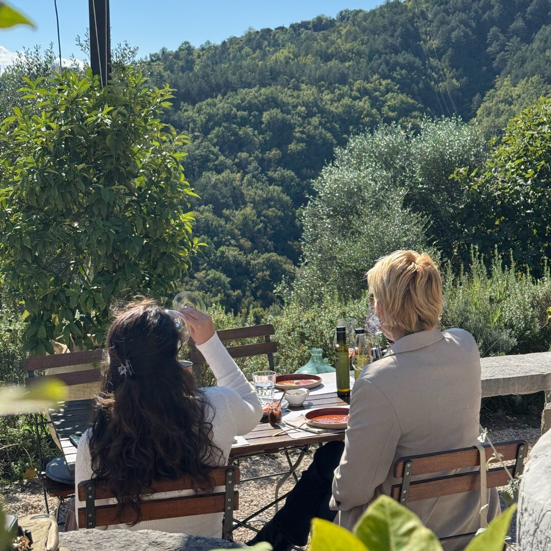 Zwei Gäste genießen eine Verkostung zu Mittag auf der Terrasse des Gutshofs Ipša, mit einer Lichterkette und Ausblicken auf ein bewaldetes Tal in Istrien. Zwei Gäste genießen eine Verkostung zu Mittag auf der Terrasse des Gutshofs Ipša, mit einer Lichterkette und Ausblicken auf ein bewaldetes Tal in Istrien.
