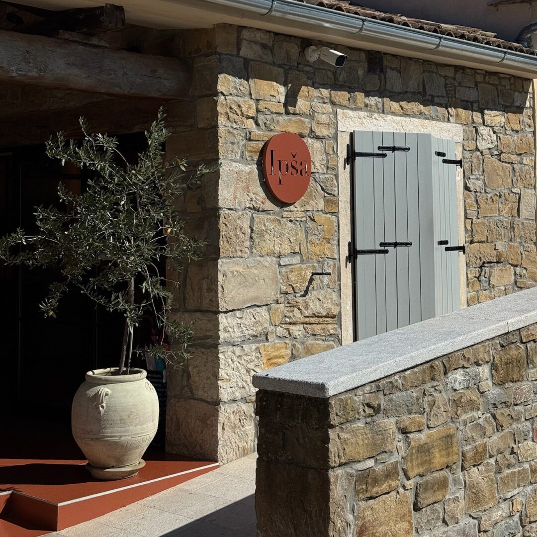 Steinfassade auf dem Gutshof Ipša mit einer roten runden Tafel, blauen Fensterläden und einem Olivenbaum im Topf. Steinfassade auf dem Gutshof Ipša mit einer roten runden Tafel, blauen Fensterläden und einem Olivenbaum im Topf.