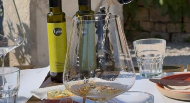 Nahaufnahme eines Verkostungstisches auf den Gutshof Ipša, mit nativen Olivenölen extra, Brot, Käse, Fleischwaren, Marmeladen und Weißwein, der eingeschenkt wird.