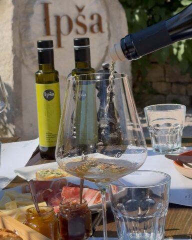 Nahaufnahme eines Verkostungstisches auf den Gutshof Ipša, mit nativen Olivenölen extra, Brot, Käse, Fleischwaren, Marmeladen und Weißwein, der eingeschenkt wird.