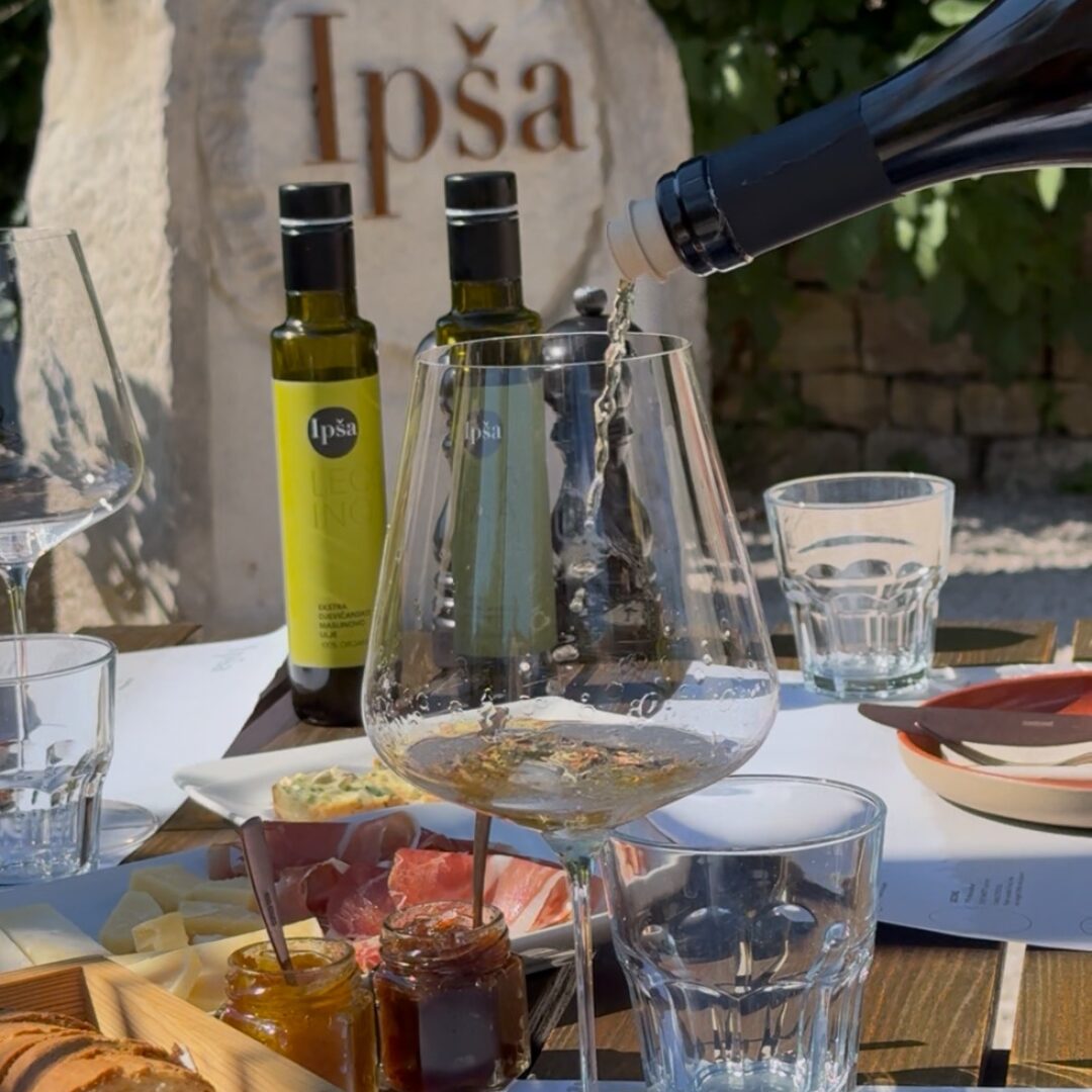 Nahaufnahme eines Verkostungstisches auf den Gutshof Ipša, mit nativen Olivenölen extra, Brot, Käse, Fleischwaren, Marmeladen und Weißwein, der eingeschenkt wird. Nahaufnahme eines Verkostungstisches auf den Gutshof Ipša, mit nativen Olivenölen extra, Brot, Käse, Fleischwaren, Marmeladen und Weißwein, der eingeschenkt wird.