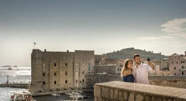 Ein Paar schießt ein Selfie an den alten Stadtmauern von Dubrovnik, mit dem schimmernden alten Hafen und der Johannesfestung im Hintergrund.