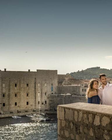 Couple taking a selfie on Dubrovnik’s ancient city walls with the Old Port and St. John Fortress shimmering in the background.