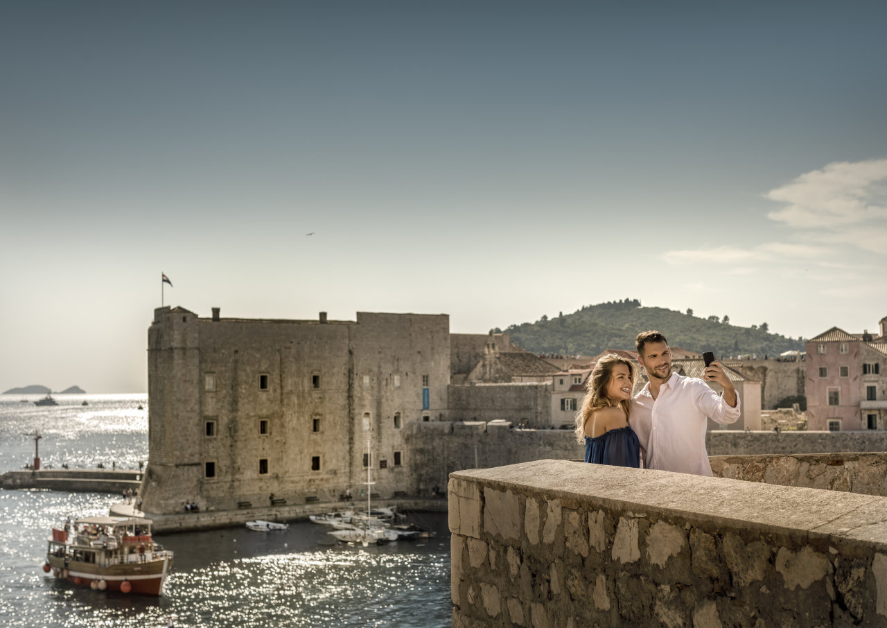 Couple taking a selfie on Dubrovnik&rsquo;s ancient city walls with the Old Port and St. John Fortress shimmering in the background. 