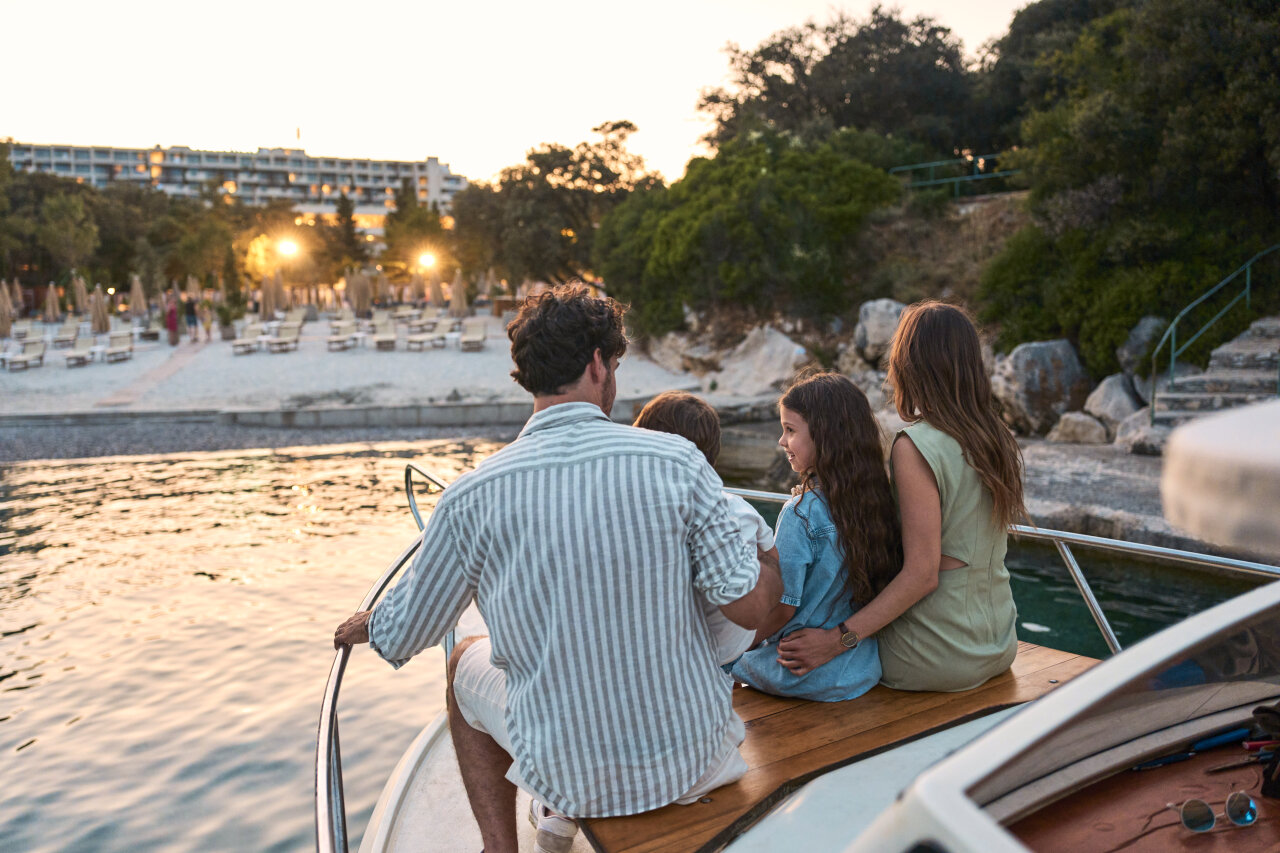 Family of four sitting on the stern of a small boat at sunset, approaching a sandy beach with loungers