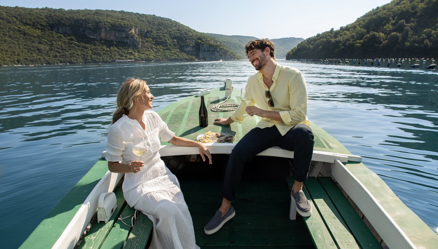 A couple enjoys a wine and seafood tasting on a small boat surrounded by calm, clear waters and lush green hills. A couple enjoys a wine and seafood tasting on a small boat surrounded by calm, clear waters and lush green hills.