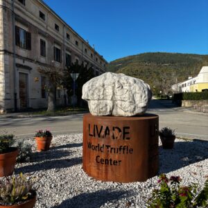 Stone truffle sculpture on a rusted steel pedestal reading ‘LIVADE World Truffle Centre,’ in a village square with historic façades and a wooded hill under a clear blue sky.