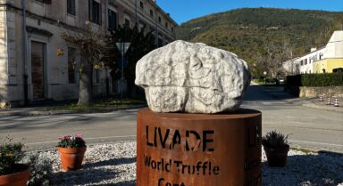 Stone truffle sculpture on a rusted steel pedestal reading ‘LIVADE World Truffle Centre,’ in a village square with historic façades and a wooded hill under a clear blue sky.