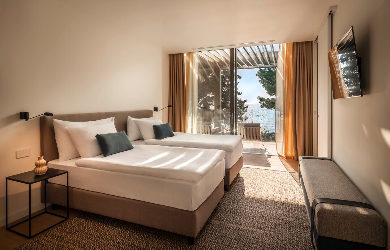 Modernes Zweibettzimmer in warmen Neutralt&ouml;nen, mit Balkonsitzpl&auml;tzen und raumhohen Glasfenstern mit Meerblick im Pical. 