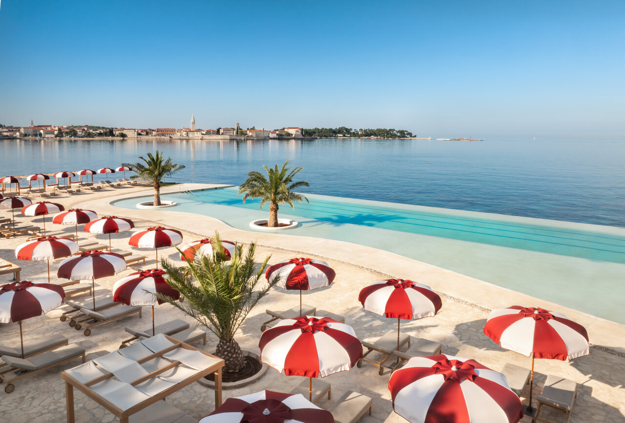Infinity-Pool-Terrasse mit rot-wei&szlig;en Sonnenschirmen und Palmeninseln, mit Blick auf die ruhige Adria und die Skyline der Altstadt von Poreč im sp&auml;ten Herbst. 