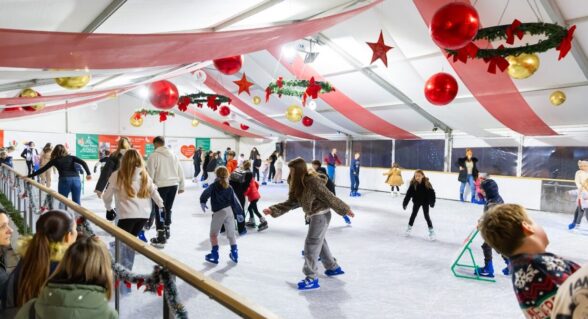 People of various ages are ice skating inside a festive indoor rink.