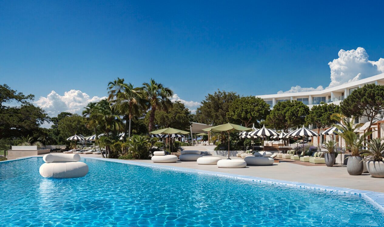 Sonniger Au&szlig;enpool im Pical, umrahmt von Palmen, Sonnenschirmen und niedrigen runden Liegen, mit dem Resort im Hintergrund. 