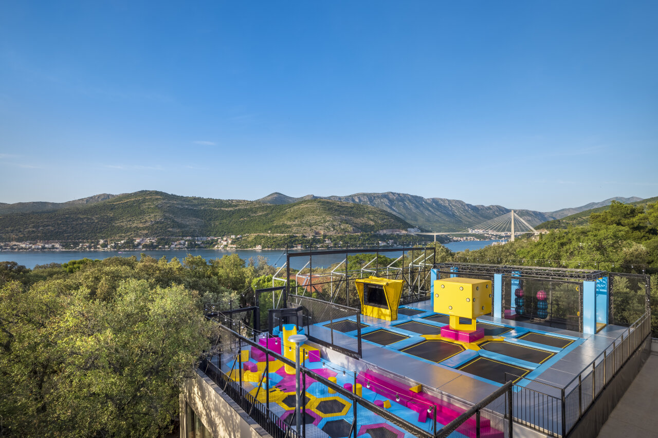 Colourful outdoor adventure park with trampolines and geometric play structures set on a hillside overlooking a bay in Dubrovnik. Ein farbenfroher Abenteuerpark im Freien, mit Trampolinen und geometrischen Erlebniswelten an einem Berghang mit Blick auf eine Bucht in Dubrovnik.
