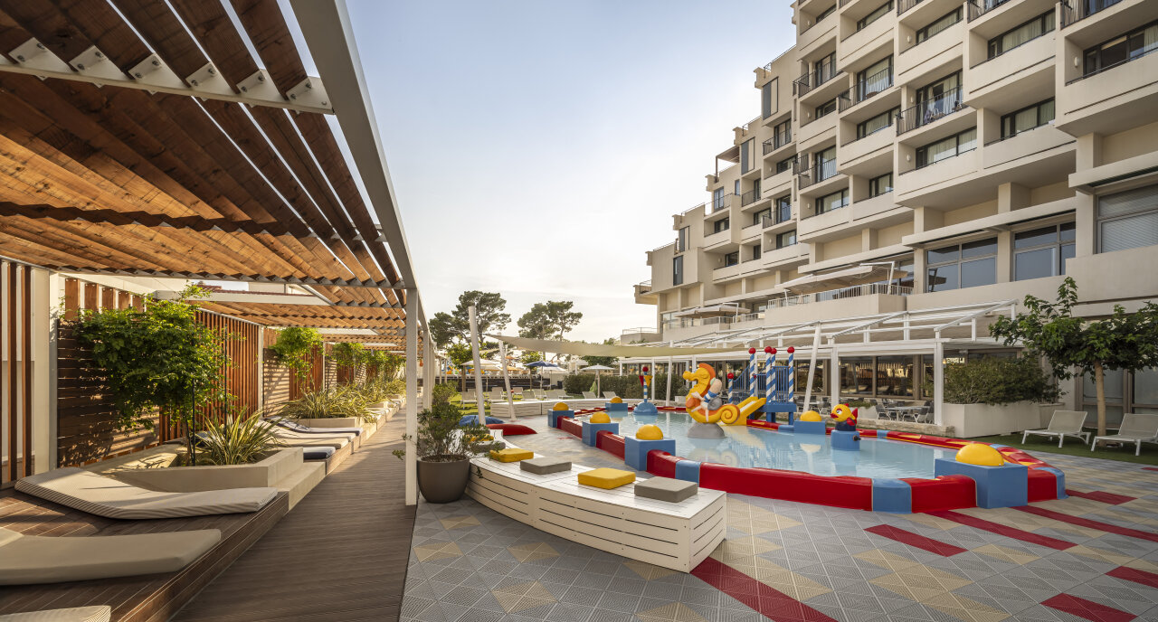 A modern family hotel courtyard at Valamar Meteor Makarska with a colourful children’s splash pool, loungers, and shaded seating areas. Ein moderner Innenhof des Familienhotels Valamar Meteor Makarska mit farbenfrohen Planschbecken, Sonnenliegen uns Sitzbereichen im Schatten.