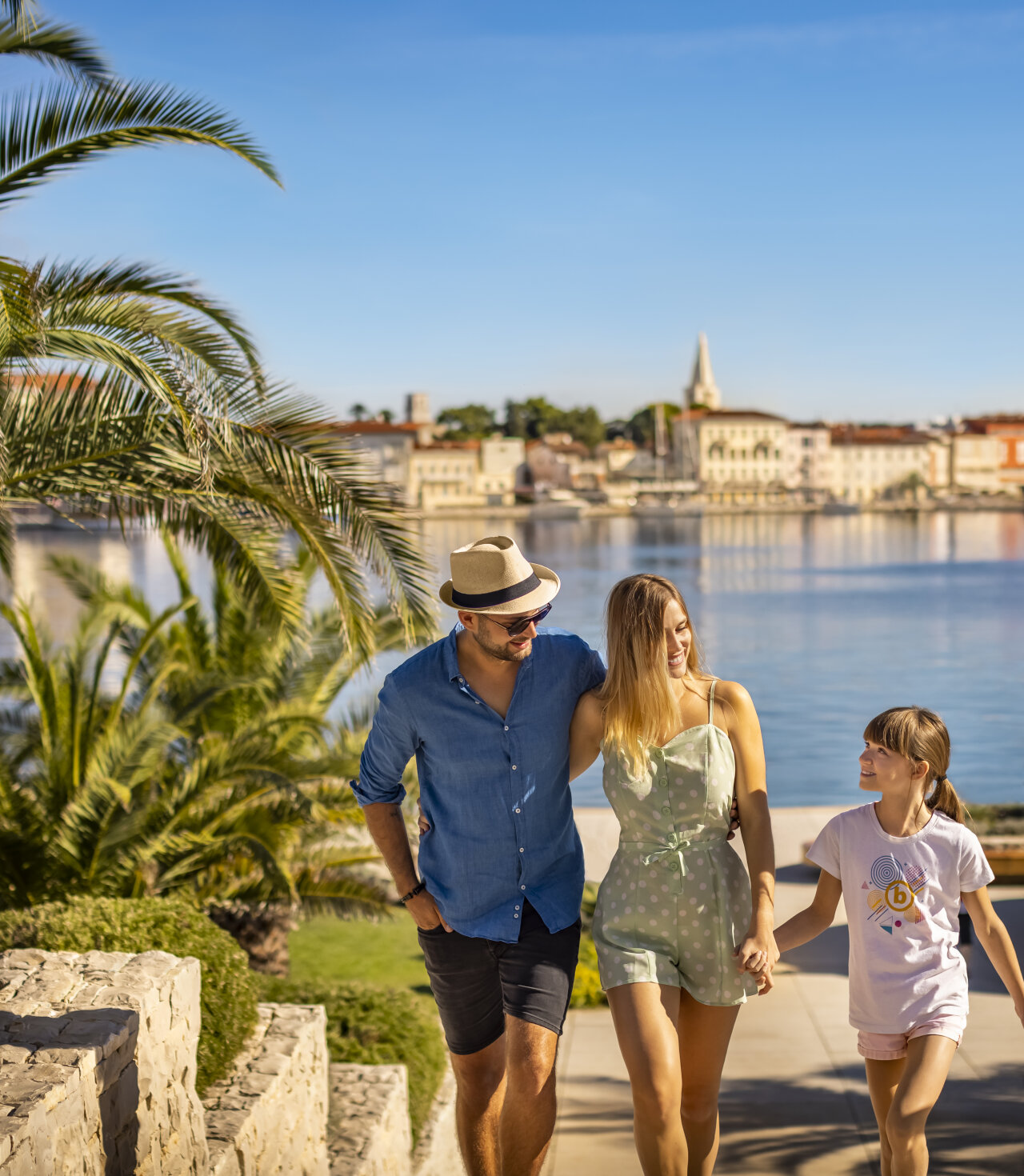 Eine Familie spaziert h&auml;ndchenhaltend auf einer K&uuml;stenpromenade mit Palmen und Poreč im Hintergrund. 