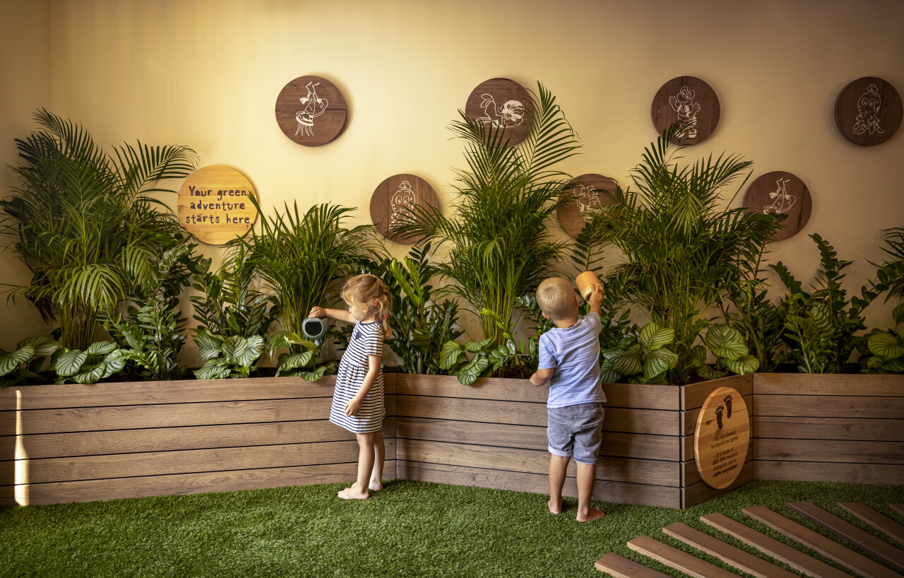 Two small children standing barefoot on artificial grass, watering a row of lush green plants in a wooden planter. Zwei kleine Kinder stehen barfuß auf einem Kunstrasen und begießen üppige grüne Pflanzen in einem hölzernen Pflanztrog.