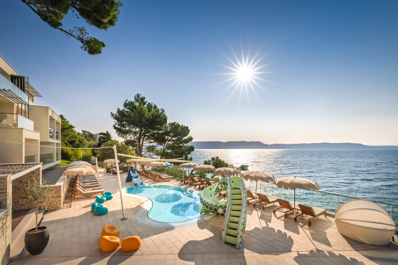 A seaside hotel pool area at Girandella Resort in Rabac with a children’s slide, sun loungers, and umbrellas overlooking the sparkling Adriatic Sea under a bright blue sky. Ein Hotelpoolbereich direkt am Meer im Girandella Resort in Rabac, mit Kinderrutsche, Sonnenliegen und Sonnenschirmen mit Blick auf die schimmernde Adria unter strahlend blauem Himmel.