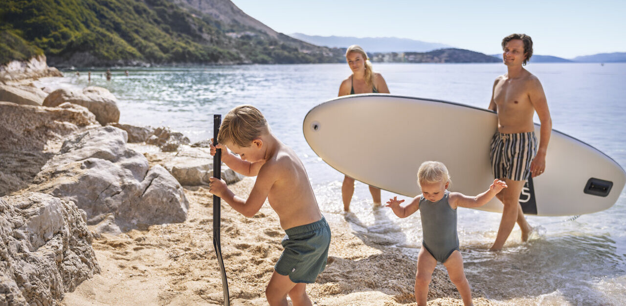 Eine Familie genie&szlig;t einen Sommertag auf einem Kiesstrand in Istrien; Kinder spielen am Meeresufer und die Erwachsenen tragen ein SUP-Board