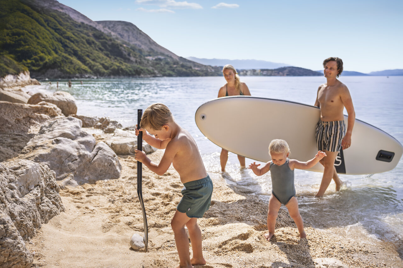 Family enjoying a summer day on a pebble beach in Istria, with children playing by the sea while adults carry a paddleboard.