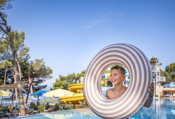 A smiling child in a swimming pool holding a large striped inflatable ring, with colourful water slides, sun umbrellas, and trees in the background under a bright blue sky.