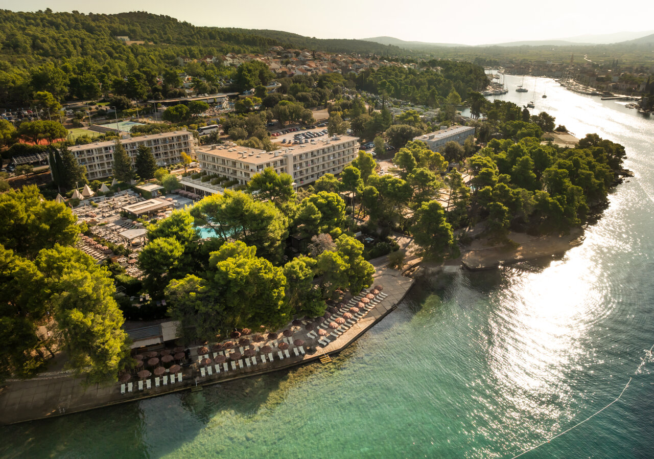 An aerial view of Stari Grad on Hvar Island, showing a seaside resort surrounded by pine trees, clear turquoise water, and a calm bay with boats and the town in the background. Luftansicht von Stari Grad auf der Insel Hvar. Zu sehen ist ein Resort direkt am Meer, umsäumt von Kiefernbäumen, klares türkisfarbenes Wasser, eine ruhige Bucht mit Booten und das Städtchen im Hintergrund.
