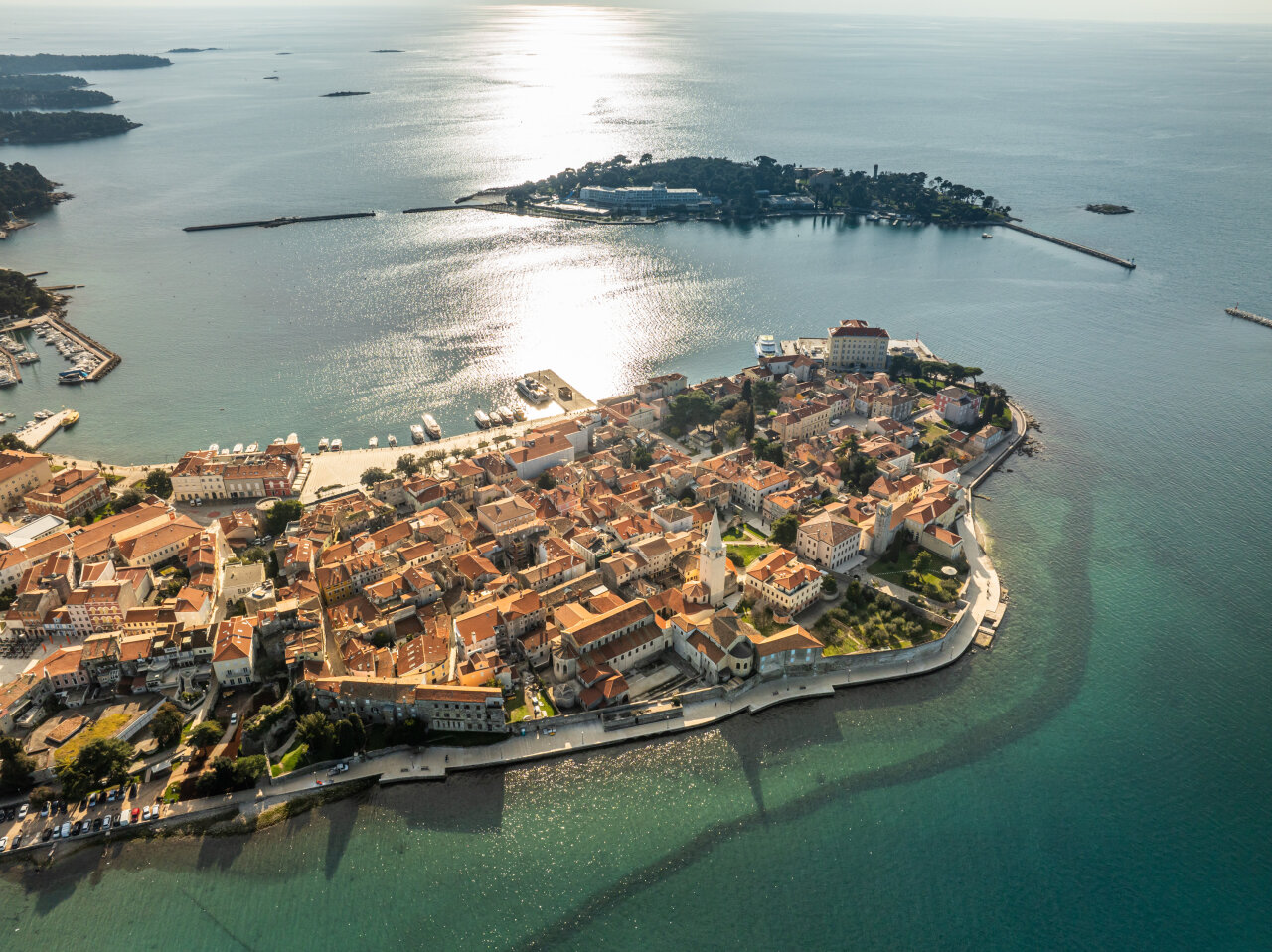 An aerial view of the historic old town of Poreč on a small peninsula, with terracotta rooftops surrounded by clear blue Adriatic waters and a nearby island in the background. Luftansicht von der historischen Altstadt von Poreč auf einer kleinen Halbinsel, mit Terracotta-Dächern, umgeben vom klaren blauen Wasser der Adria und eine nahe gelegene Insel im Hintergrund.