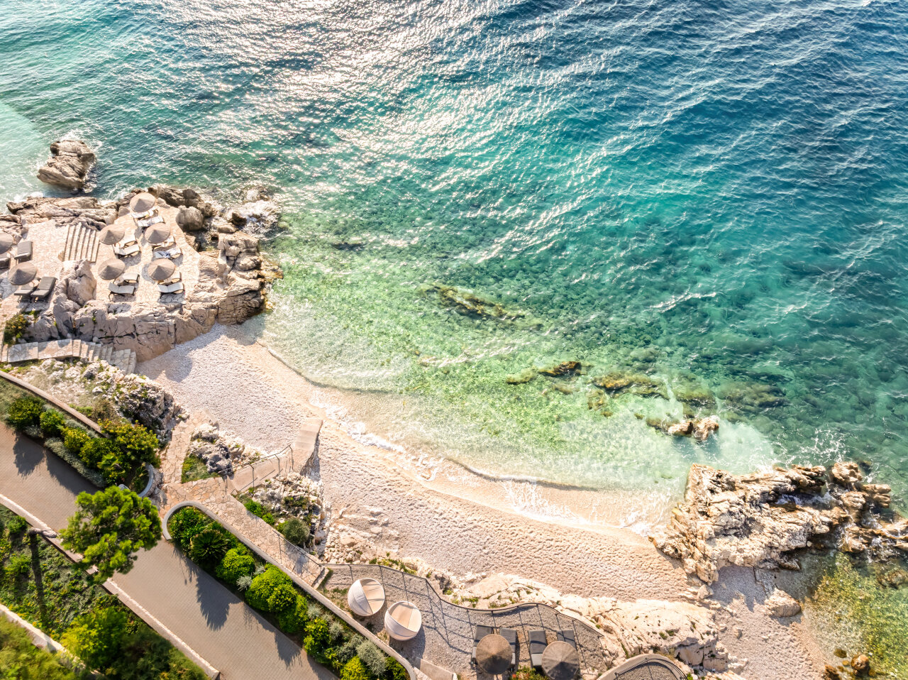 Aerial view of a sandy beach with turquoise Adriatic Sea, stone sun terraces, and coastal pathways in Rabac. 