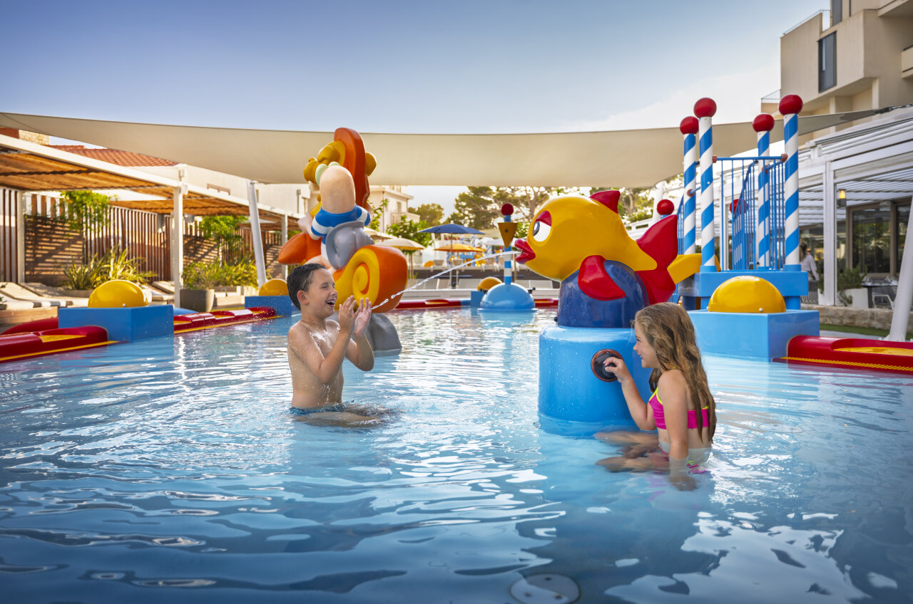 Two children play in a shallow splash pool surrounded by bright fish-shaped water features and spray fountains.