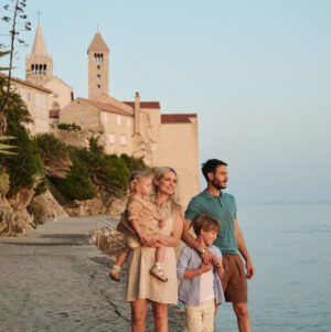 Zwei Erwachsene und zwei Kinder stehen auf einer K&uuml;stenpromenade nahe bei steinernen Geb&auml;uden und den Kircht&uuml;rmen der Stadt Rab, den Blick auf das ruhige Wasser gerichtet.